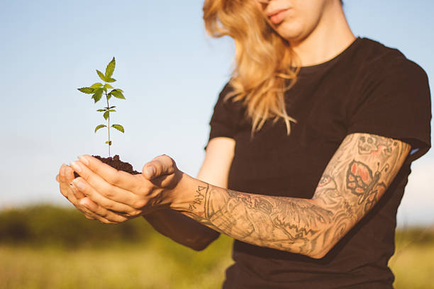 Person with tattooed arms cradles sapling; no tattoo ink visible.