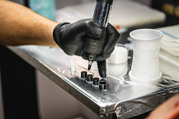 Gloved hand dips tattoo machine into caps of black tattoo ink on foil-covered tray.