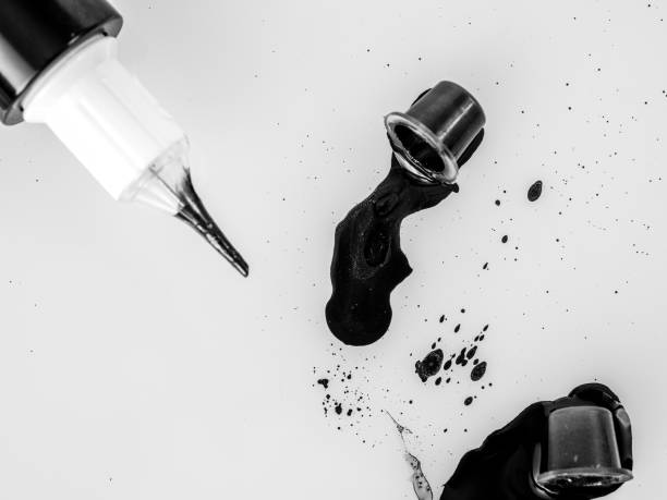 Black tattoo ink spilled from cap beside needle tube on white surface.
