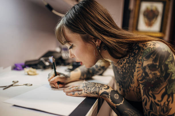 A tattoo artist with arm tattoos sketches a design; tattoo ink is in use.
