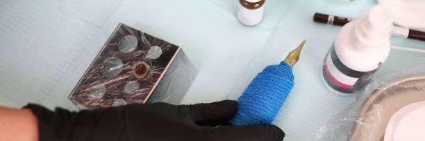 Gloved hand holds tattoo machine, with tattoo ink and supplies on a sterile table.